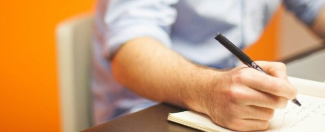 Man at desk using laptop with book and pen
